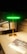 A modern, sleek lamp with a golden base and stem, topped by a glowing green horizontal light strip. It stands on a wooden tabletop next to a brochure displaying similar lighting designs. There is a reflection of the lamp on a glass surface behind it, enhancing the room's contemporary and stylish decor.