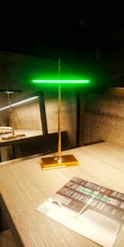 A modern, sleek lamp with a golden base and stem, topped by a glowing green horizontal light strip. It stands on a wooden tabletop next to a brochure displaying similar lighting designs. There is a reflection of the lamp on a glass surface behind it, enhancing the room's contemporary and stylish decor.