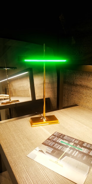 A modern, sleek lamp with a golden base and stem, topped by a glowing green horizontal light strip. It stands on a wooden tabletop next to a brochure displaying similar lighting designs. There is a reflection of the lamp on a glass surface behind it, enhancing the room