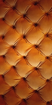 A detailed close-up of a tufted leather surface featuring a grid of cushioned, diamond-shaped patterns. The leather appears soft and plush, with stitching forming the intersections, creating a luxurious and ornate texture.