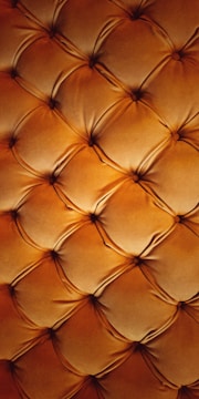 A detailed close-up of a tufted leather surface featuring a grid of cushioned, diamond-shaped patterns. The leather appears soft and plush, with stitching forming the intersections, creating a luxurious and ornate texture.