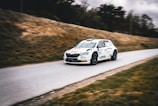 A rally car speeds down a narrow road, surrounded by grassy hills. The vehicle is emblazoned with decals and sponsorship logos, hinting at a professional motorsport event. The background is slightly blurred, emphasizing the car's motion.