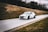 A rally car speeds down a narrow road, surrounded by grassy hills. The vehicle is emblazoned with decals and sponsorship logos, hinting at a professional motorsport event. The background is slightly blurred, emphasizing the car's motion.