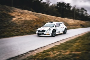 A rally car speeds down a narrow road, surrounded by grassy hills. The vehicle is emblazoned with decals and sponsorship logos, hinting at a professional motorsport event. The background is slightly blurred, emphasizing the car's motion.