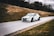 A rally car speeds down a narrow road, surrounded by grassy hills. The vehicle is emblazoned with decals and sponsorship logos, hinting at a professional motorsport event. The background is slightly blurred, emphasizing the car's motion.