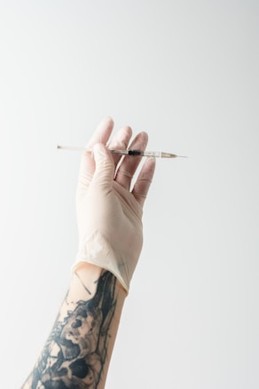 A hand wearing a latex glove holds a syringe filled with liquid. The arm is adorned with a tattoo depicting detailed artwork. The background is plain and light-colored, providing contrast to the arm and syringe.