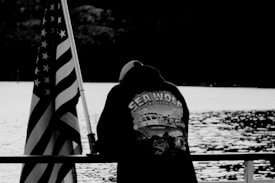 A person wearing a hooded jacket with a design featuring a ship, sea wolf, and the text 'SEA WOLF EMERYVILLE, CA.' leans on a railing by the water. Next to them is an American flag on a pole. The image is in black and white, with the water reflecting light in the background.