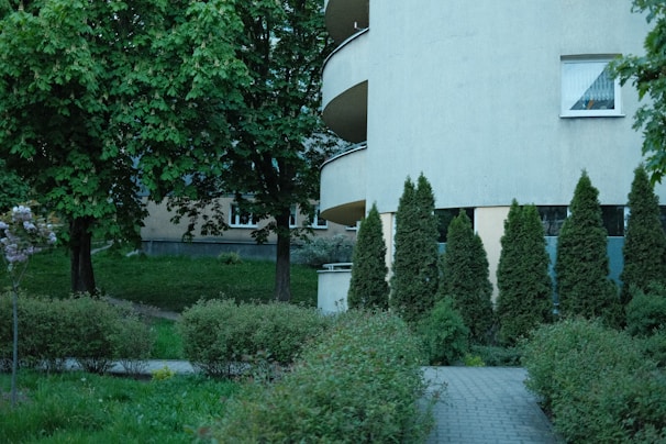A peaceful urban scene featuring a residential building with curved balconies partially obscured by tall, well-kept hedges and trees. The foreground includes a paved pathway surrounded by lush green shrubbery and grass. Large leafy trees dominate the background, providing shade and contributing to the tranquil atmosphere.