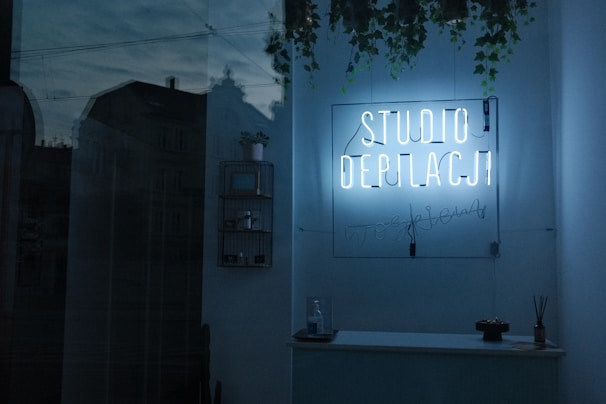 A dimly lit interior features a neon sign reading 'STUDIO DEPILACJI' on a wall adorned with hanging plants. Shelves display small items, and a counter holds a few decorations. The glass reflection reveals an exterior view with faint outlines of buildings.