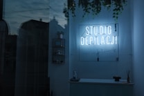 A dimly lit interior features a neon sign reading 'STUDIO DEPILACJI' on a wall adorned with hanging plants. Shelves display small items, and a counter holds a few decorations. The glass reflection reveals an exterior view with faint outlines of buildings.
