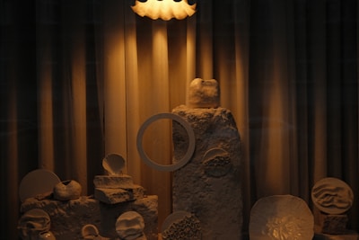 A dimly lit display featuring a variety of abstract sculptures made from natural materials. The scene includes textured stones and uniquely shaped ceramic pieces, arranged artistically on a small platform. The lighting casts warm, soft shadows across the objects, enhancing their textures and forms.