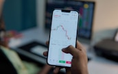 A person holds a smartphone displaying a financial or stock trading app, with detailed graphs and numerical data visible on the screen. In the background, there is a blurred computer monitor and office setting, suggesting a work environment.