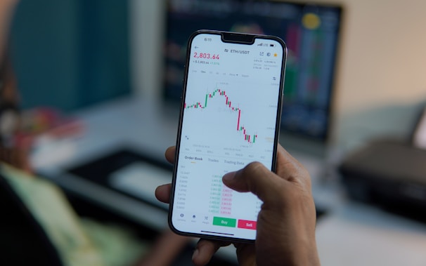 A person holds a smartphone displaying a financial or stock trading app, with detailed graphs and numerical data visible on the screen. In the background, there is a blurred computer monitor and office setting, suggesting a work environment.