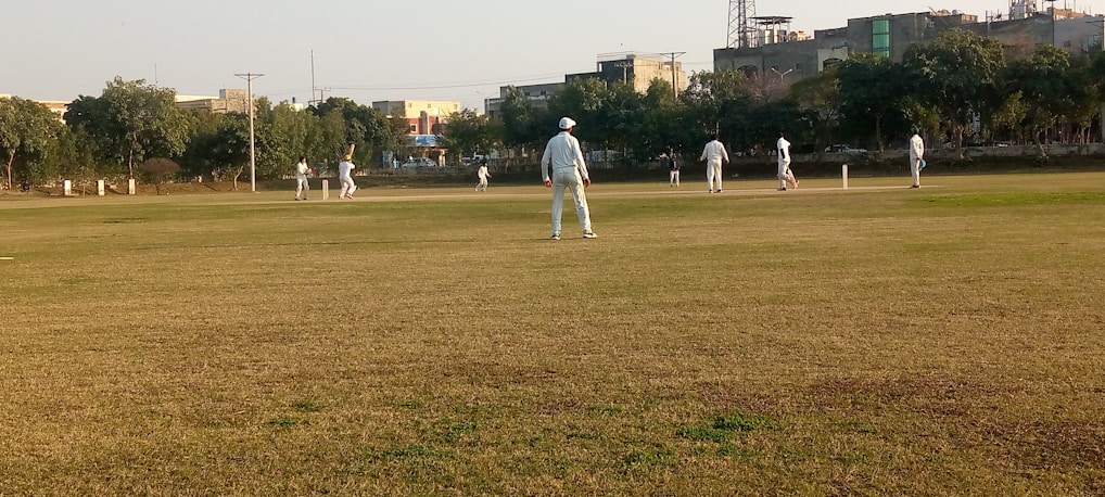 Several people are playing a cricket match on a large grassy field. The players are dressed in light-colored sports uniforms, and some of them are wearing caps. Trees and buildings can be seen in the background, indicating an urban park setting. The sky is clear, suggesting a sunny day.