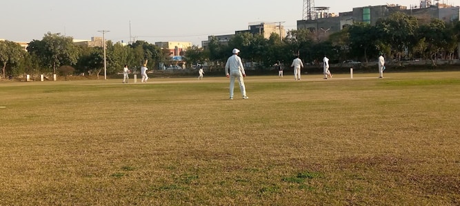 Several people are playing a cricket match on a large grassy field. The players are dressed in light-colored sports uniforms, and some of them are wearing caps. Trees and buildings can be seen in the background, indicating an urban park setting. The sky is clear, suggesting a sunny day.