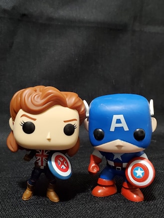 Two Funko Pop figurines are standing together against a dark background. The figurine on the left features a character with brown hair and a Union Jack design on the chest, holding a shield with a similar pattern. The figurine on the right depicts a character in a blue costume with an 'A' on the forehead and a star emblem on the chest, holding a red, white, and blue shield.
