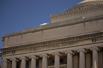 The image depicts the façade of a neoclassical building, featuring tall columns and a triangular pediment. The engraved text reads 'The Library of Columbia University'. The architecture is characterized by detailed stone carvings and inscriptions, creating a sense of grandeur.