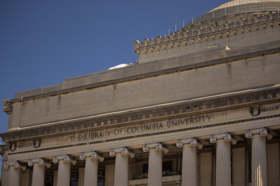 The image depicts the façade of a neoclassical building, featuring tall columns and a triangular pediment. The engraved text reads 'The Library of Columbia University'. The architecture is characterized by detailed stone carvings and inscriptions, creating a sense of grandeur.