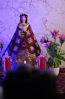 A decorative statue of a praying woman, adorned in a richly patterned maroon and gold garment. It is placed on a marble background, flanked by two red candles with faux flames and placed beside vibrant pink flowers in white pots that add a touch of liveliness.