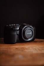 A professional digital camera sits on a wooden surface with a dark background. Its lens is missing, revealing the mount, and there are several buttons and controls visible on its body.