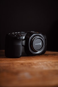 A professional digital camera sits on a wooden surface with a dark background. Its lens is missing, revealing the mount, and there are several buttons and controls visible on its body.