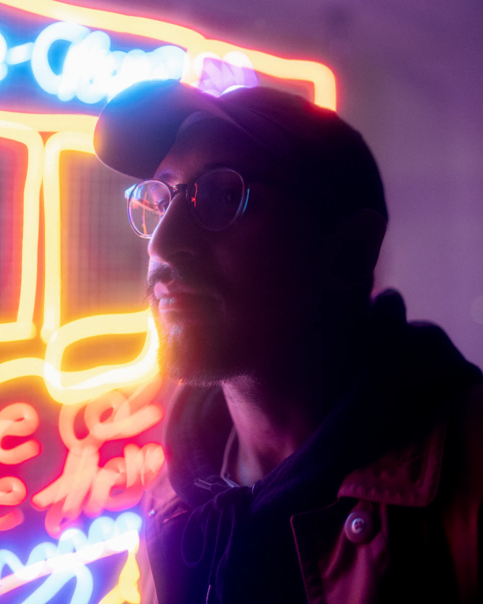 The image shows a man with glasses and a black baseball cap, wearing a brown jacket, standing against a purple background illuminated by neon lights. The man's face is turned to the side, suggesting he might be looking off into the distance or observing something in his peripheral vision.