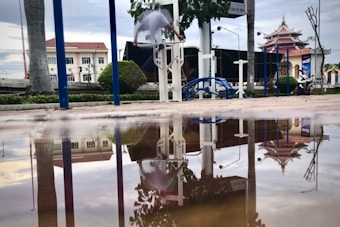 A person is engaged in a physical workout, using outdoor gym equipment in an urban setting. The scene includes a large puddle in the foreground, which reflects the buildings, trees, and the gym equipment. The background shows a temple-like structure alongside modern buildings, emphasizing a blend of traditional and contemporary architecture.