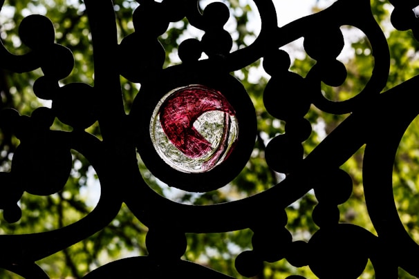 Intricate black metalwork with a small, colorful stained glass window in the center. The glass features a vibrant red and clear pattern, surrounded by lush green foliage in the background, slightly out of focus.