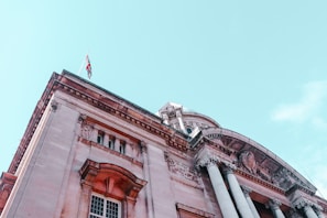 A grand, neoclassical-style building with large columns and intricate architectural details. A flag is seen waving atop the structure against a clear sky.
