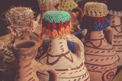A collection of decorative clay jugs adorned with colorful knitted covers on their lids. The jugs feature intricate painted patterns in earthy tones, and each lid is uniquely designed with vibrant threads, adding a touch of handmade artistry.