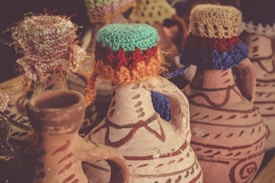 A collection of decorative clay jugs adorned with colorful knitted covers on their lids. The jugs feature intricate painted patterns in earthy tones, and each lid is uniquely designed with vibrant threads, adding a touch of handmade artistry.