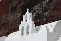 A white church with bells and crosses stands out against a rocky, earthy-colored cliff background. The architectural style is minimalistic, highlighting clean lines and contrasting colors.
