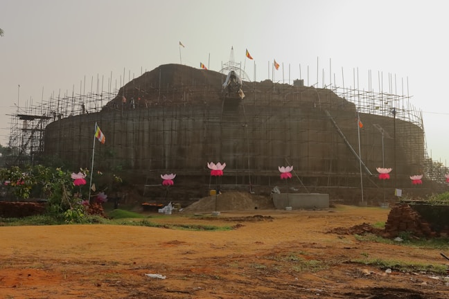A large structure under construction is surrounded by scaffolding with several pink lotus-shaped decorations attached. Flags are flying on tall poles, and the ground is a mixture of soil and grass. There are some construction materials visible near the base.