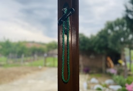 A strand of green beads or prayer beads hangs from a dark-colored hook attached to a wooden surface. The background is a blurred outdoor scene with trees, a garden, and a cloudy sky.