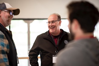 A group of people engaging in a cheerful conversation indoors. The focus is on a man wearing glasses, a black jacket, and a lanyard, who is smiling broadly. The setting appears casual and light-hearted, with a soft, defocused background.