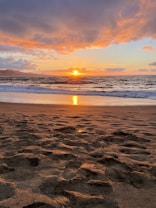The image captures a serene sunset over the ocean, with vibrant colors illuminating the sky. Wispy clouds are spread across the horizon, reflecting the warm orange and pink hues of the setting sun. In the foreground, sandy footprints lead to the waves gently lapping at the shore, creating a peaceful atmosphere.