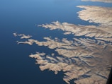 An aerial view of a sprawling landmass with intricate shapes surrounded by a deep blue body of water. The land appears dry, with a mix of brown and beige tones, and is intersected by narrow channels of water.
