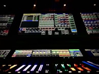 A complex control panel with multiple monitors displaying various technical interfaces, featuring grids and buttons in different colors. The bottom part shows illuminated controls, possibly indicating a sound or lighting board.