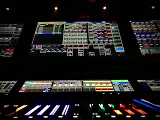 A complex control panel with multiple monitors displaying various technical interfaces, featuring grids and buttons in different colors. The bottom part shows illuminated controls, possibly indicating a sound or lighting board.