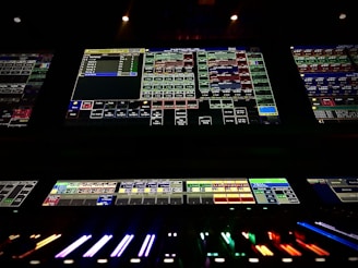 A complex control panel with multiple monitors displaying various technical interfaces, featuring grids and buttons in different colors. The bottom part shows illuminated controls, possibly indicating a sound or lighting board.