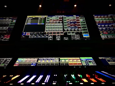 A complex control panel with multiple monitors displaying various technical interfaces, featuring grids and buttons in different colors. The bottom part shows illuminated controls, possibly indicating a sound or lighting board.