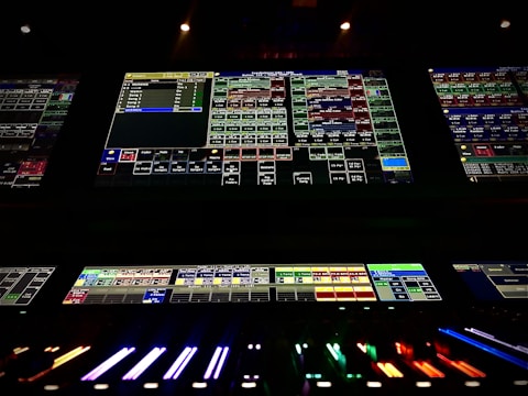 A complex control panel with multiple monitors displaying various technical interfaces, featuring grids and buttons in different colors. The bottom part shows illuminated controls, possibly indicating a sound or lighting board.
