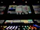 A complex control panel with multiple monitors displaying various technical interfaces, featuring grids and buttons in different colors. The bottom part shows illuminated controls, possibly indicating a sound or lighting board.