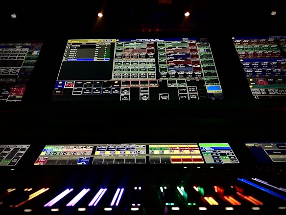 A complex control panel with multiple monitors displaying various technical interfaces, featuring grids and buttons in different colors. The bottom part shows illuminated controls, possibly indicating a sound or lighting board.