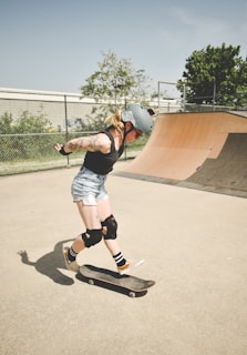 A person wearing a helmet and protective knee pads is actively skateboarding at a skate park. The skateboard is in mid-air as the person prepares to perform a trick. The scene is set in a concrete skate area with a wooden ramp and a fenced backdrop. The sunny day and clear sky highlight the vibrant outdoor atmosphere.