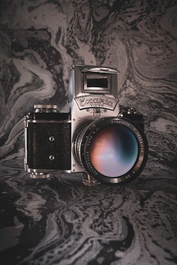 A vintage camera with a silver metallic and black body is prominently displayed. The lens shows a reflection with a gradient of colors. The background features a textured, swirling marble-like pattern in shades of gray and black, adding an artistic and classic vibe to the image.