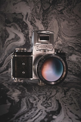 A vintage camera with a silver metallic and black body is prominently displayed. The lens shows a reflection with a gradient of colors. The background features a textured, swirling marble-like pattern in shades of gray and black, adding an artistic and classic vibe to the image.