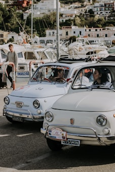 Two vintage Fiat cars are parked side by side in a marina area. The cars have an Italian club sticker on the windshields, and there are boats and buildings in the background. A person is walking nearby and another person is seated inside one of the cars.