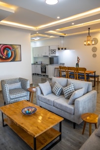 A modern living room with a cozy, stylish design featuring a grey sofa and armchair with patterned cushions. A wooden coffee table with a decorative bowl sits in the center. The room is decorated with a colorful abstract painting and elegant ceiling lights. The open-plan layout leads into a kitchen area with white cabinets and a dining area with wooden chairs.