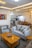 A modern living room with a cozy, stylish design featuring a grey sofa and armchair with patterned cushions. A wooden coffee table with a decorative bowl sits in the center. The room is decorated with a colorful abstract painting and elegant ceiling lights. The open-plan layout leads into a kitchen area with white cabinets and a dining area with wooden chairs.
